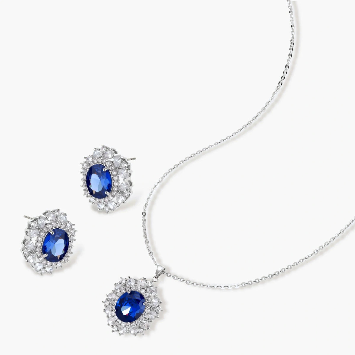 White gold plated jewelry set with blue cubic zirconia centers surrounded by clear crystal halos on cream background.